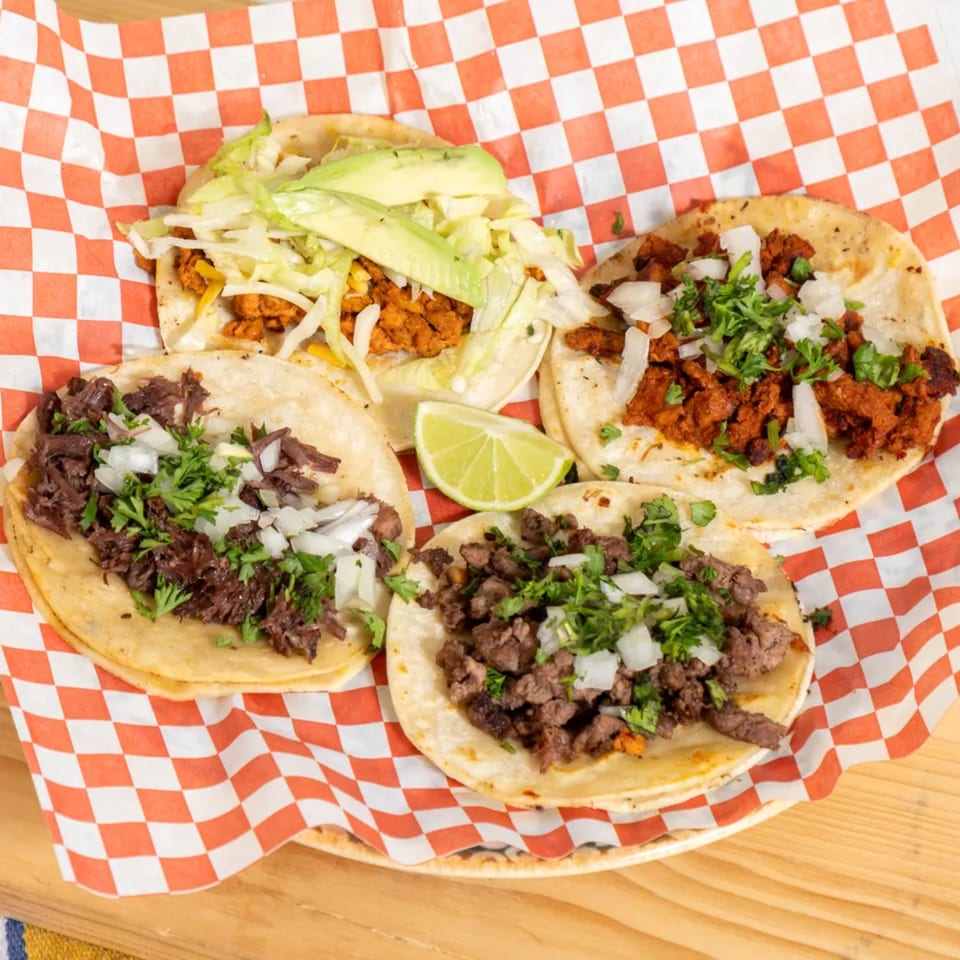 Four diverse tacos with various fillings, cilantro, onions, and avocado, served on red and white checkered paper.